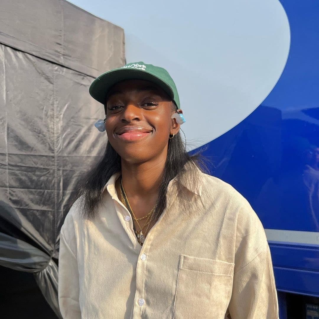 A person wearing a beige shirt and green baseball hat stands in front of a blue backdrop.